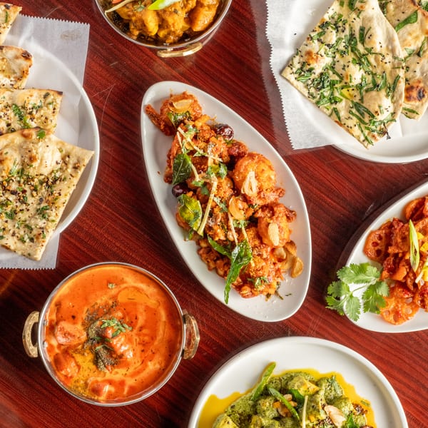 An overhead view of various Indian dishes, including naan, curries, and fried appetizers, on a wooden table.