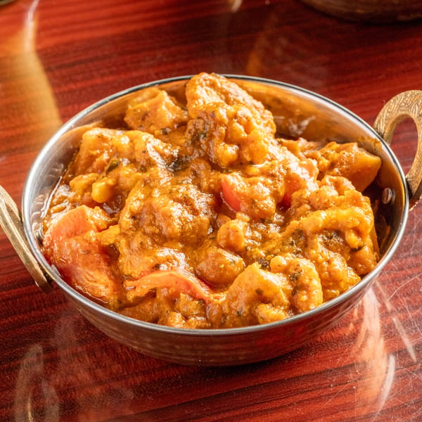 A metal bowl filled with a rich orange curry, featuring shrimp and vegetables, on a wooden table.