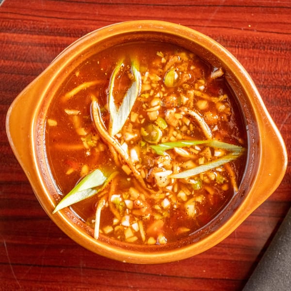 Overhead view of a bowl of hot and sour soup with scallions and spices on a wooden table.