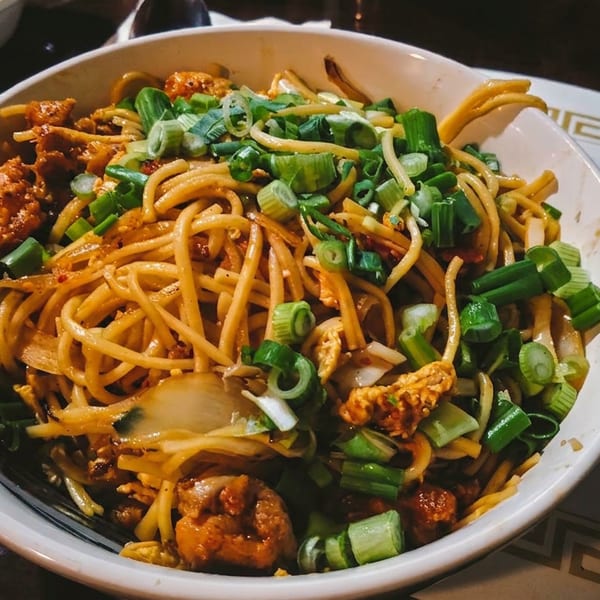 A large bowl of savory stir-fried noodles with chicken, green onions, and a fork.